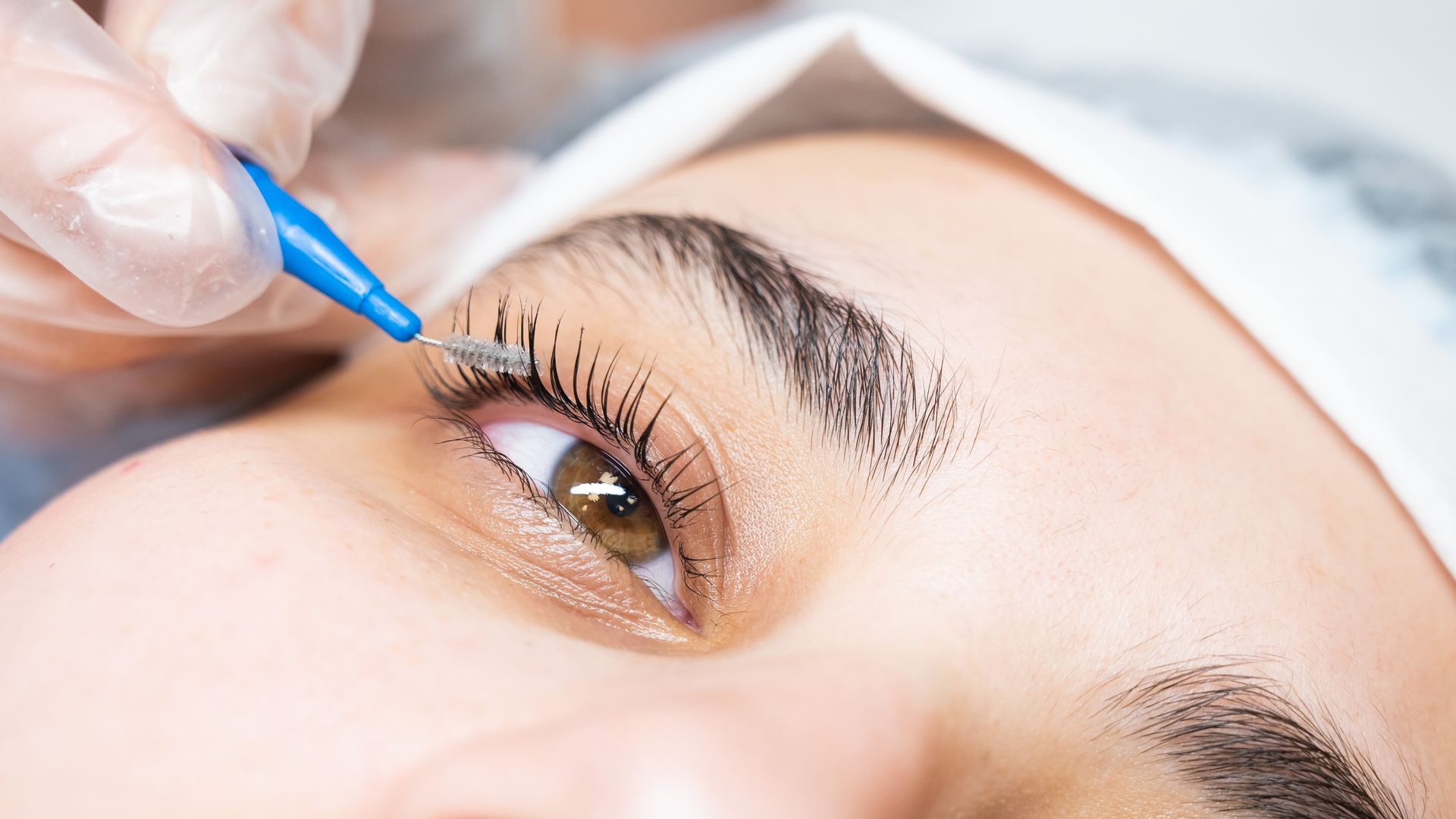 Close-up of an eyebrow being shaped with blue tweezers near a person’s eye