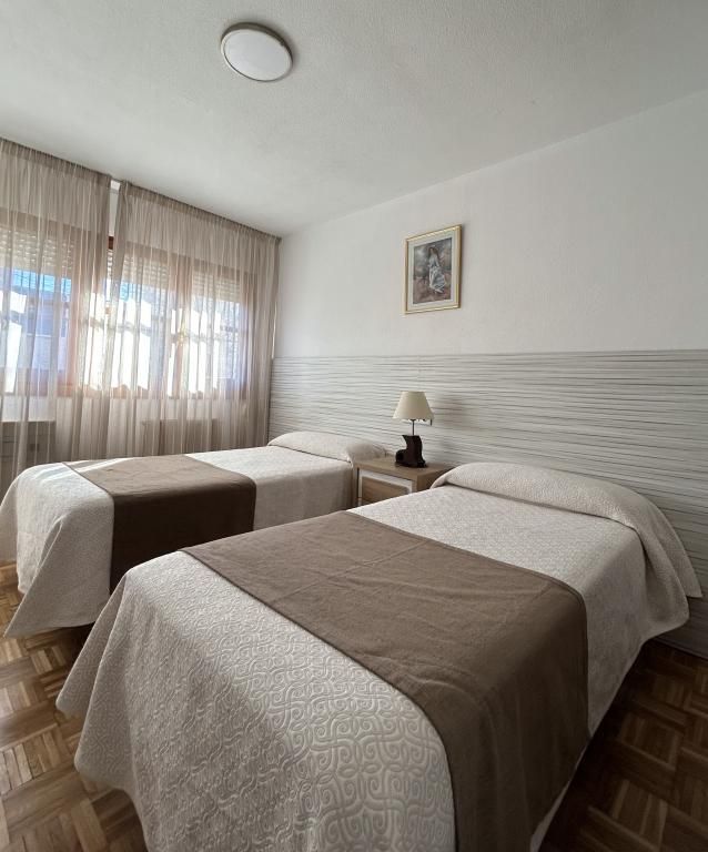 Dos camas individuales en una habitación luminosa de color beige con ventana, cortinas y una pequeña mesita de noche.