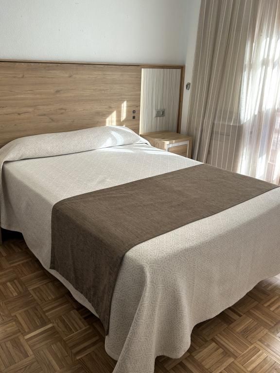 Cama bien hecha con ropa de cama blanca y marrón en un dormitorio luminoso, junto a cortinas transparentes.