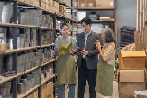 Three people discussing inventory in a warehouse aisle beside stacked shelves and boxes.