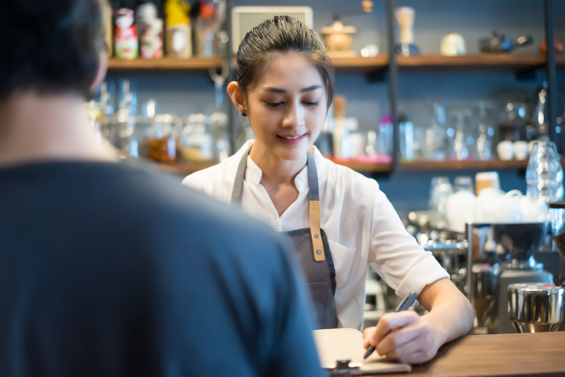 Barista in a white shirt serving coffee at a café counter, with shelves of bottles in the background