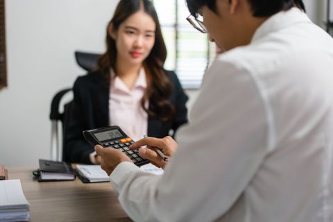 Two people in an office discussing finances while one uses a calculator at a desk