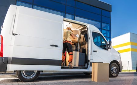White delivery van with open side door and boxes outside, parked beside a blue building