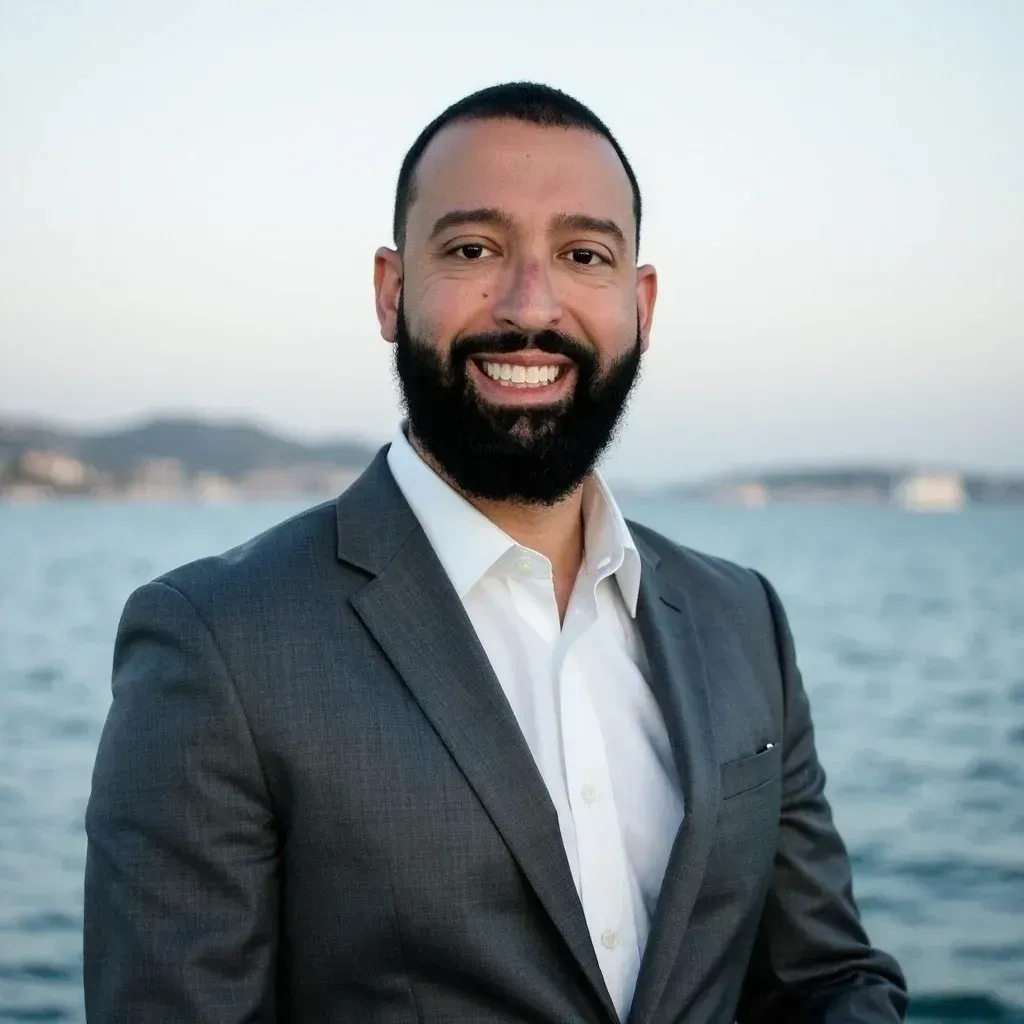 Smiling man in a gray suit by the water with boats in the background