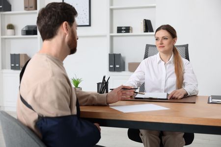 Two people in a bright office meeting across a desk, one holding papers and talking to the other.