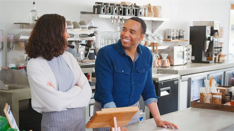 Two people talking at a bright café counter, smiling with coffee equipment in the background.