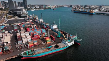 Harbor with moored cargo ships beside stacked shipping containers and waterfront buildings