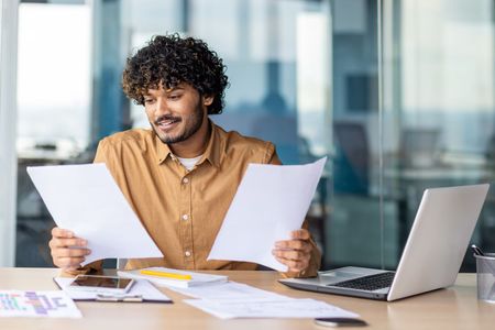 Man reviewing two documents at a desk with a laptop and papers in a bright office