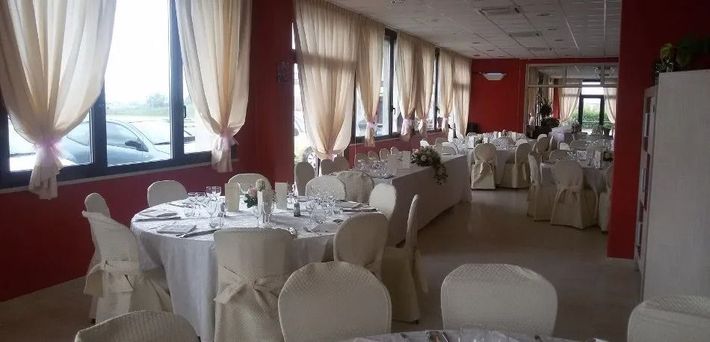 Luminosa sala da pranzo con tavoli apparecchiati con tovaglie bianche, sedie coperte e ampie finestre con tende.