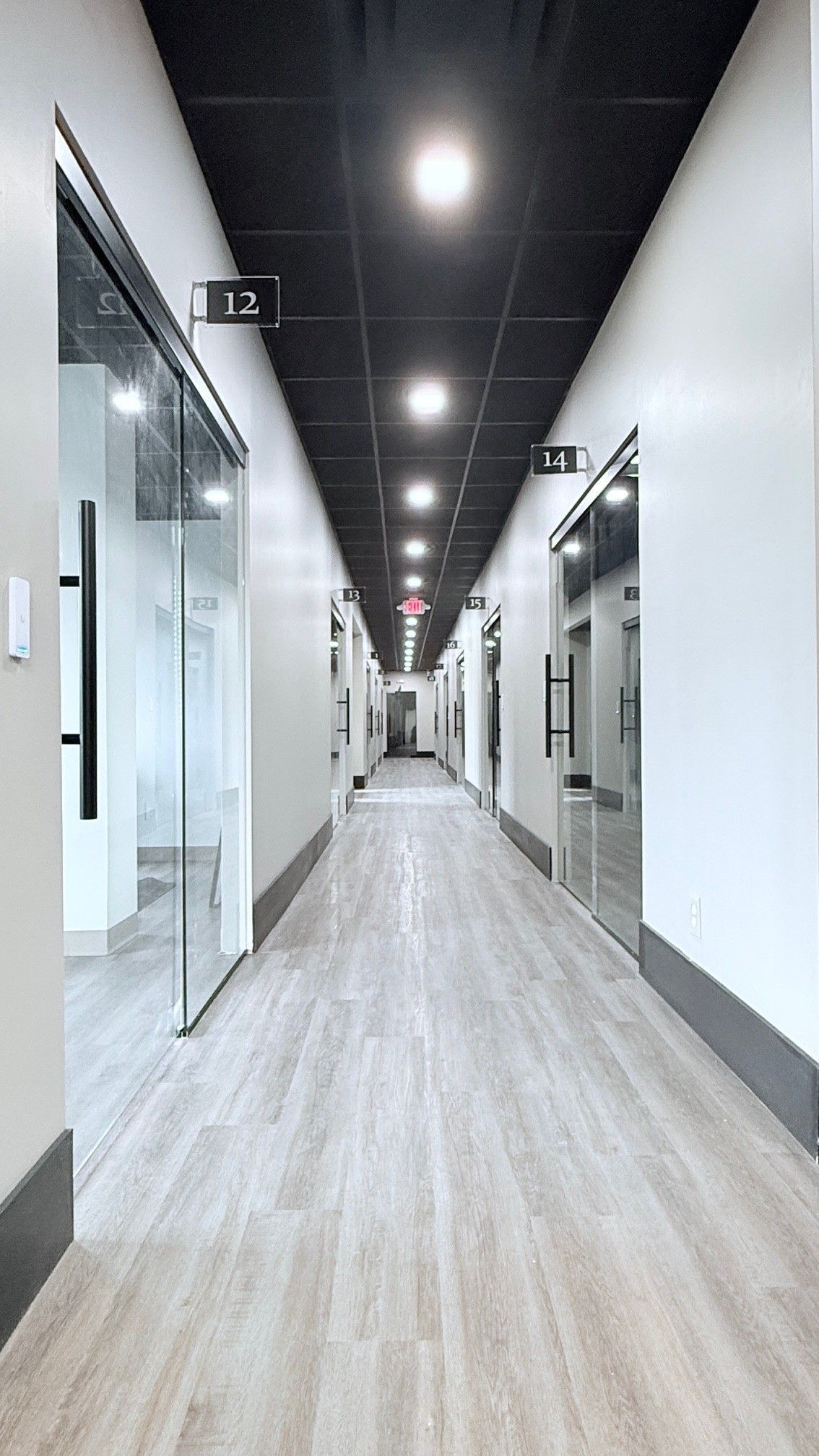 Long hallway with gray flooring, white walls, glass-walled offices, and a black ceiling with recessed lighting.