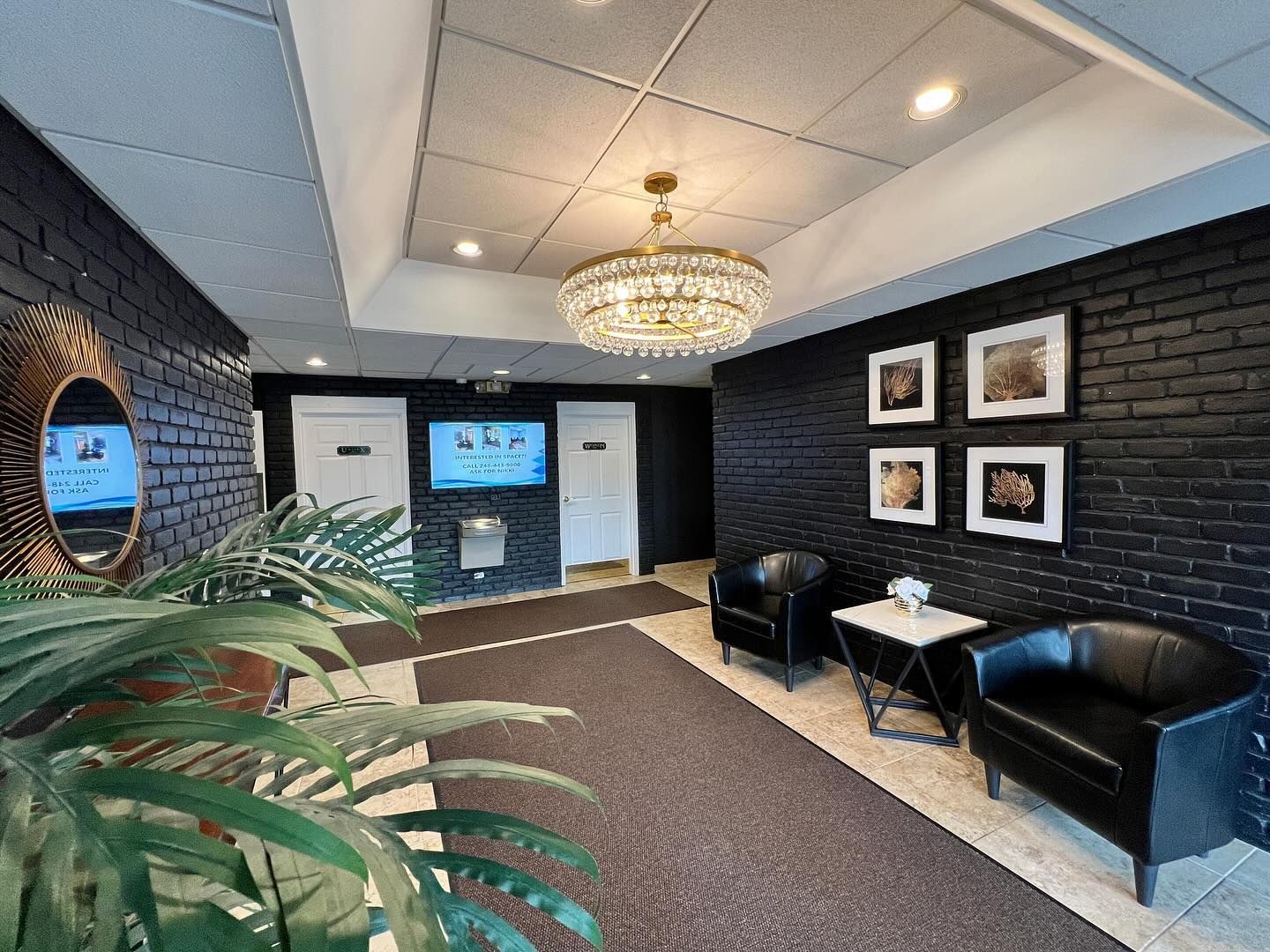 Lobby with black brick walls, chandelier, two black armchairs, artwork, mirror, and plant.