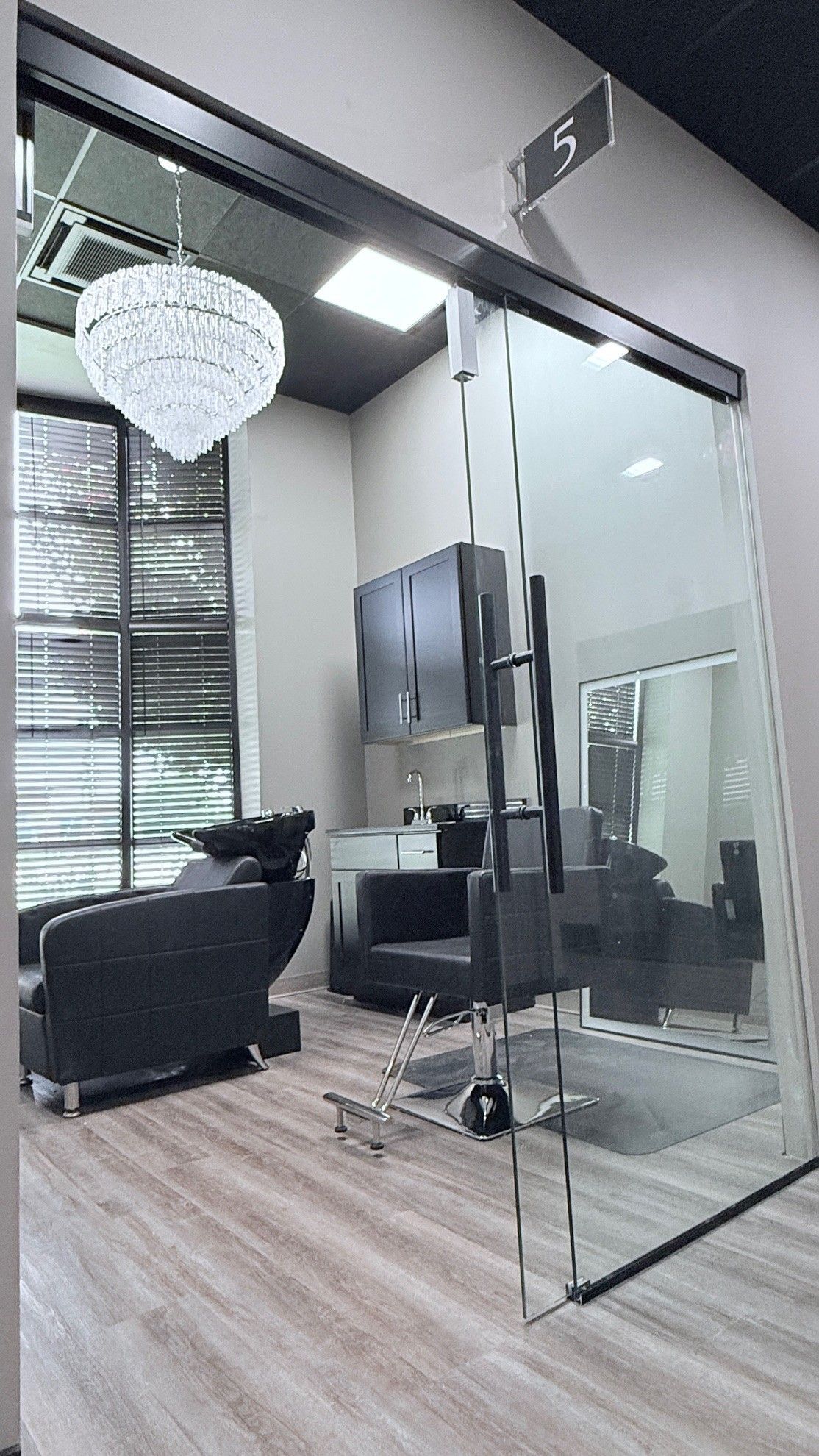 Hair salon interior: glass door, crystal chandelier, black chairs, light wood floor.