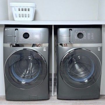 Two gray washing machines side-by-side in a laundry room with a shelf above and a laundry basket.
