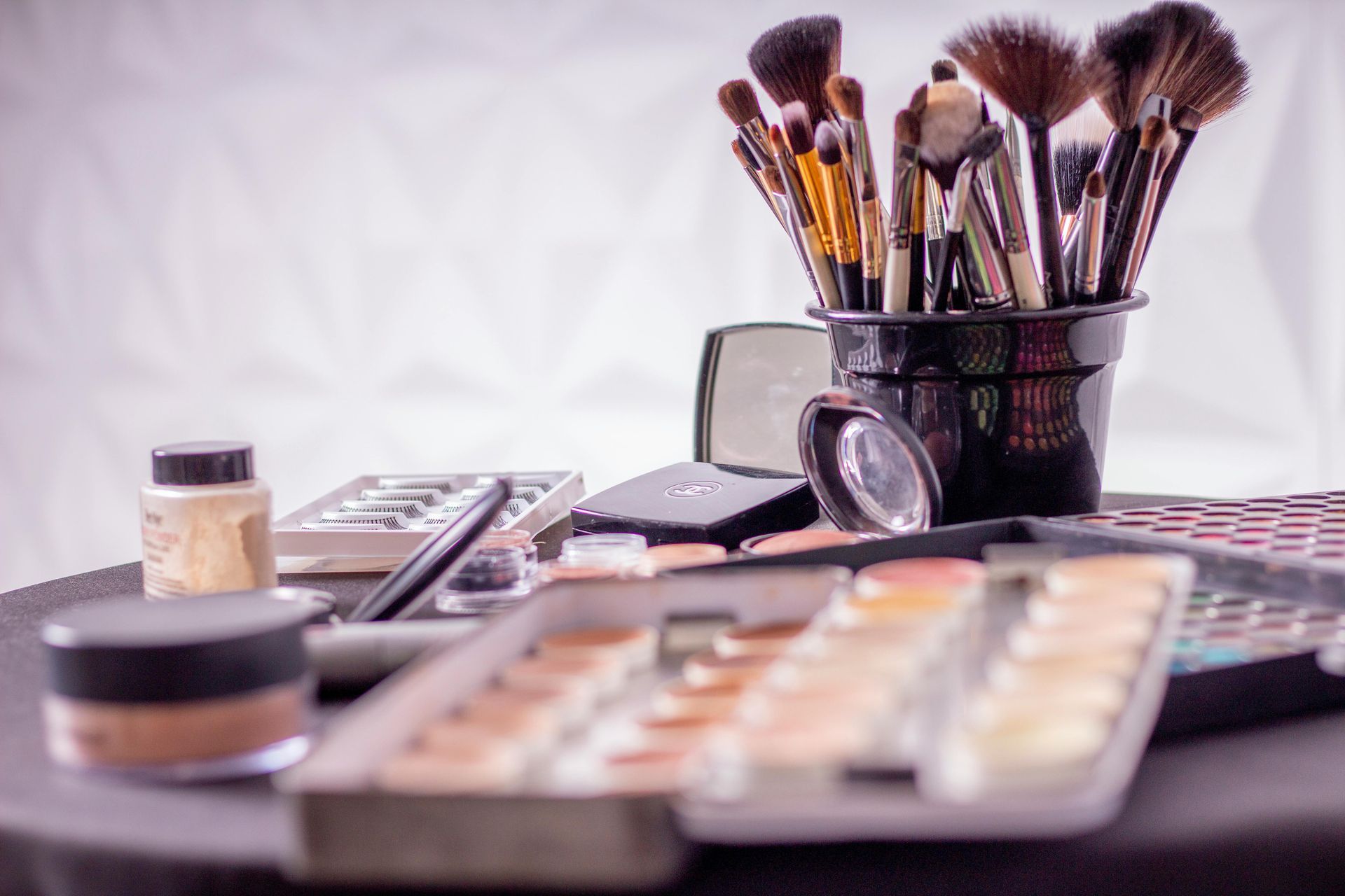 Makeup brushes in a black holder, with palettes, foundation, and other makeup products on a table.