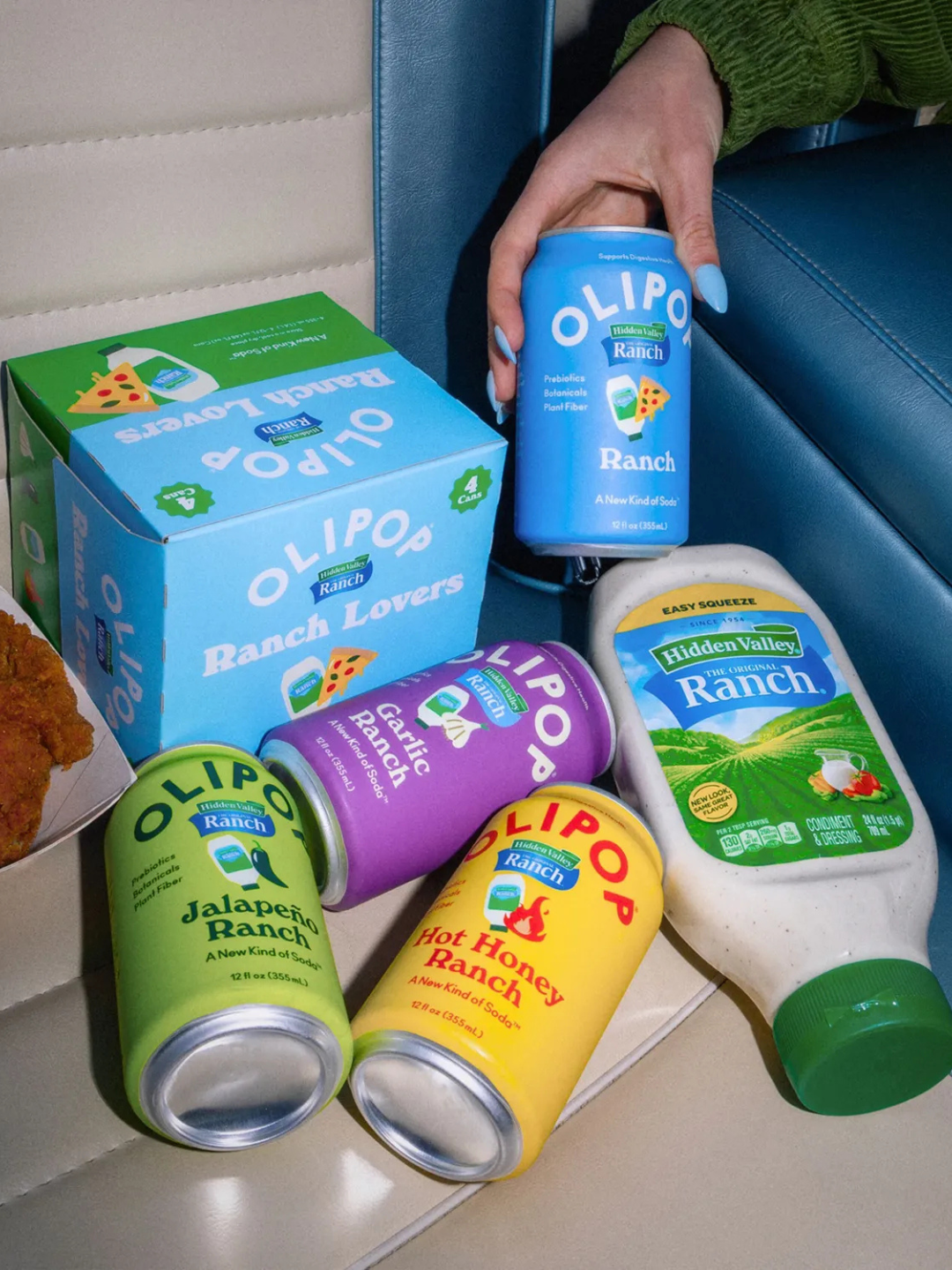 A hand holds a blue Olipop soda can, surrounded by other flavor cans and a bottle of Hidden Valley Ranch on a car seat.