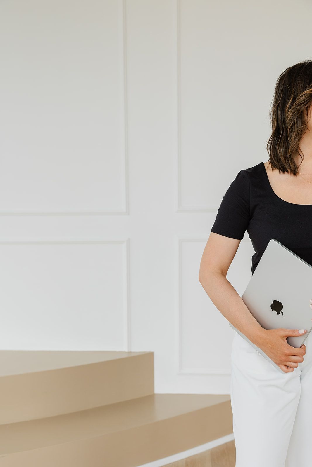 Woman holding a laptop, standing by beige stairs in a white room