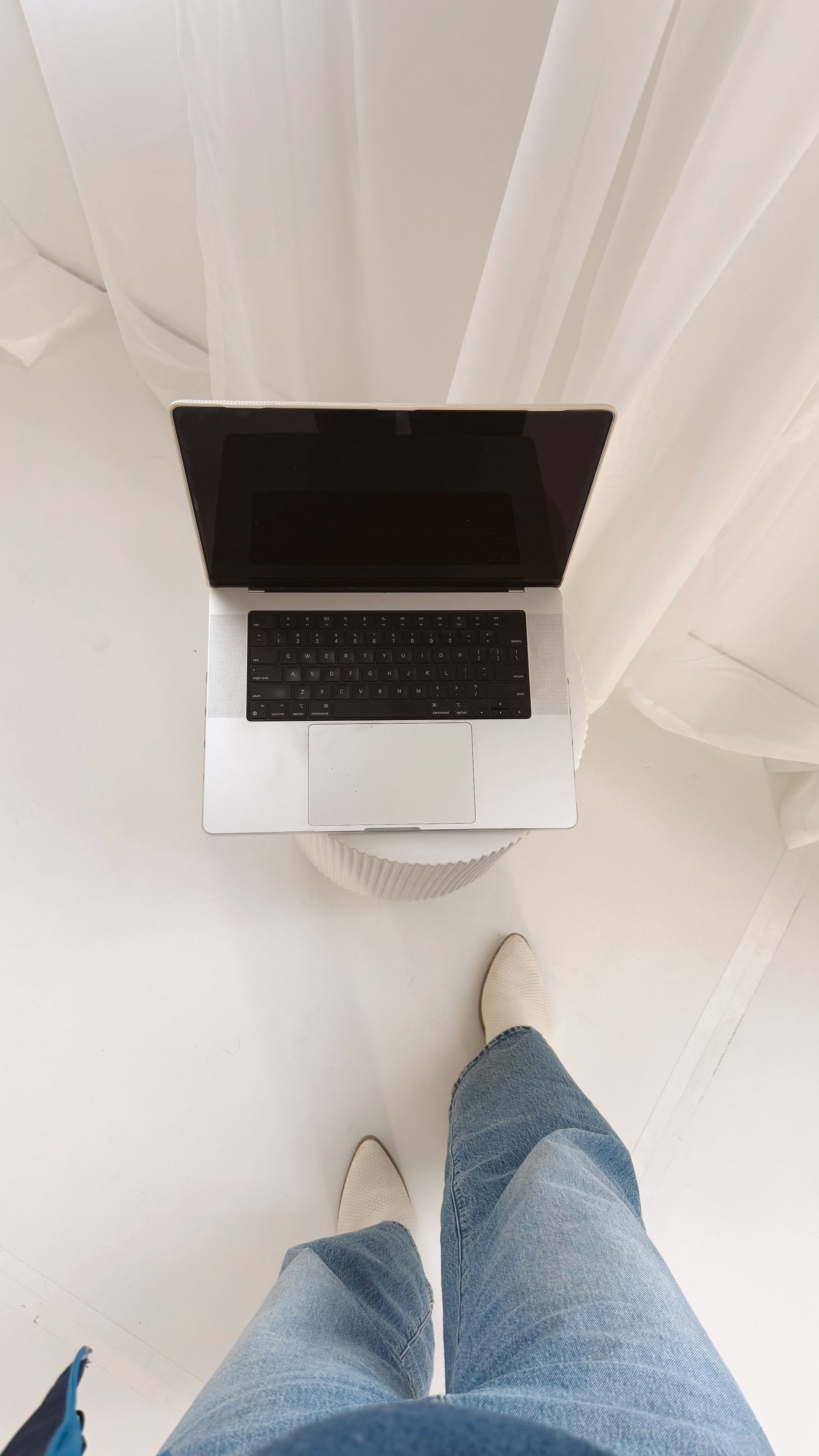 Person standing near a laptop on a white surface, wearing jeans and shoes. Bright white backdrop.