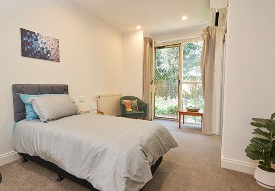 Bedroom with single bed, small table, chair, and open doorway to greenery.