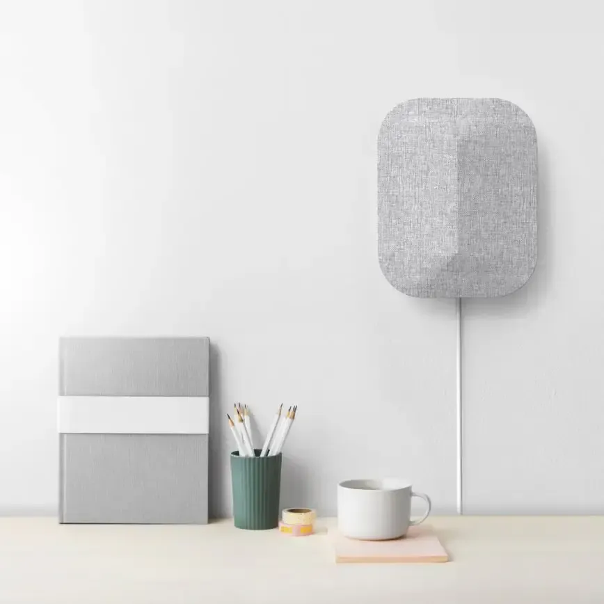 A gray fabric speaker mounted on a white wall above a desk with stationery and a mug.