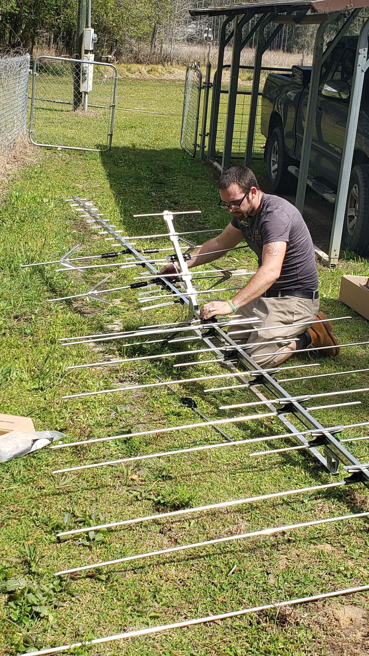Man assembling a silver outdoor TV antenna on green grass. Carport and fence in the background.