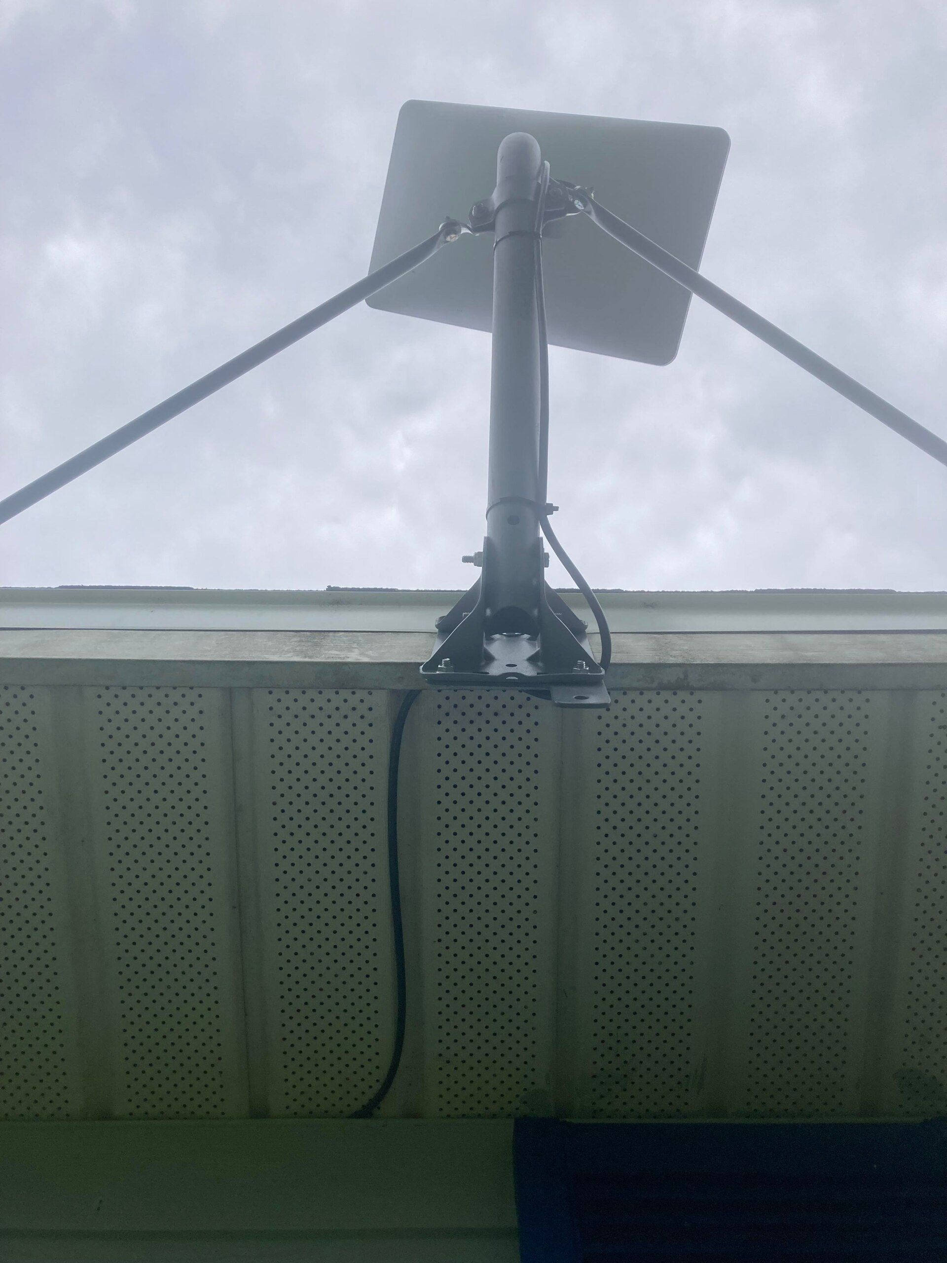 A satellite dish mounted on a house roof, supported by metal beams. Black cable hangs down. Overcast sky.