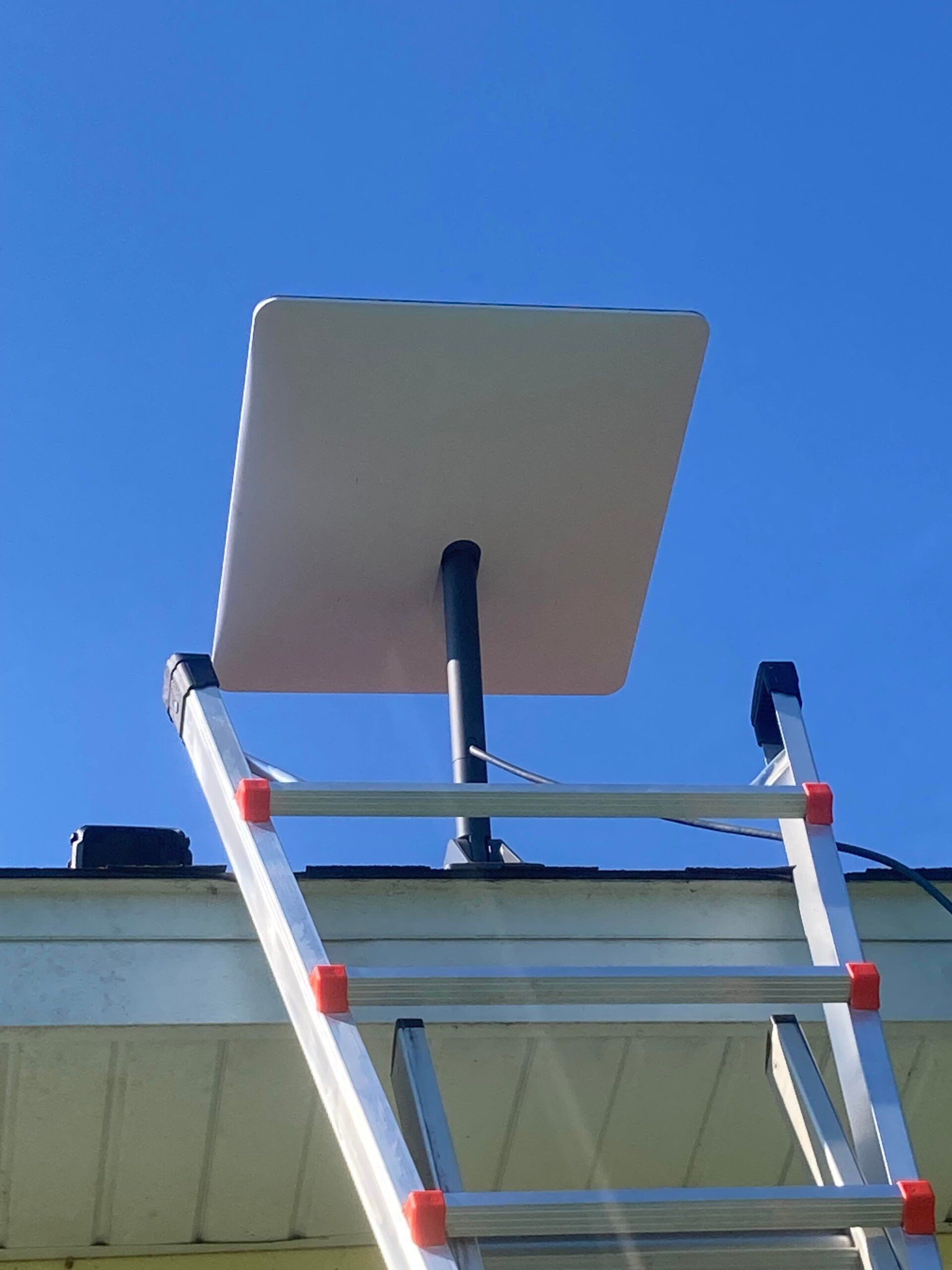 White rectangular satellite dish on roof, mounted on black pole. Ladder angled up, against a blue sky.