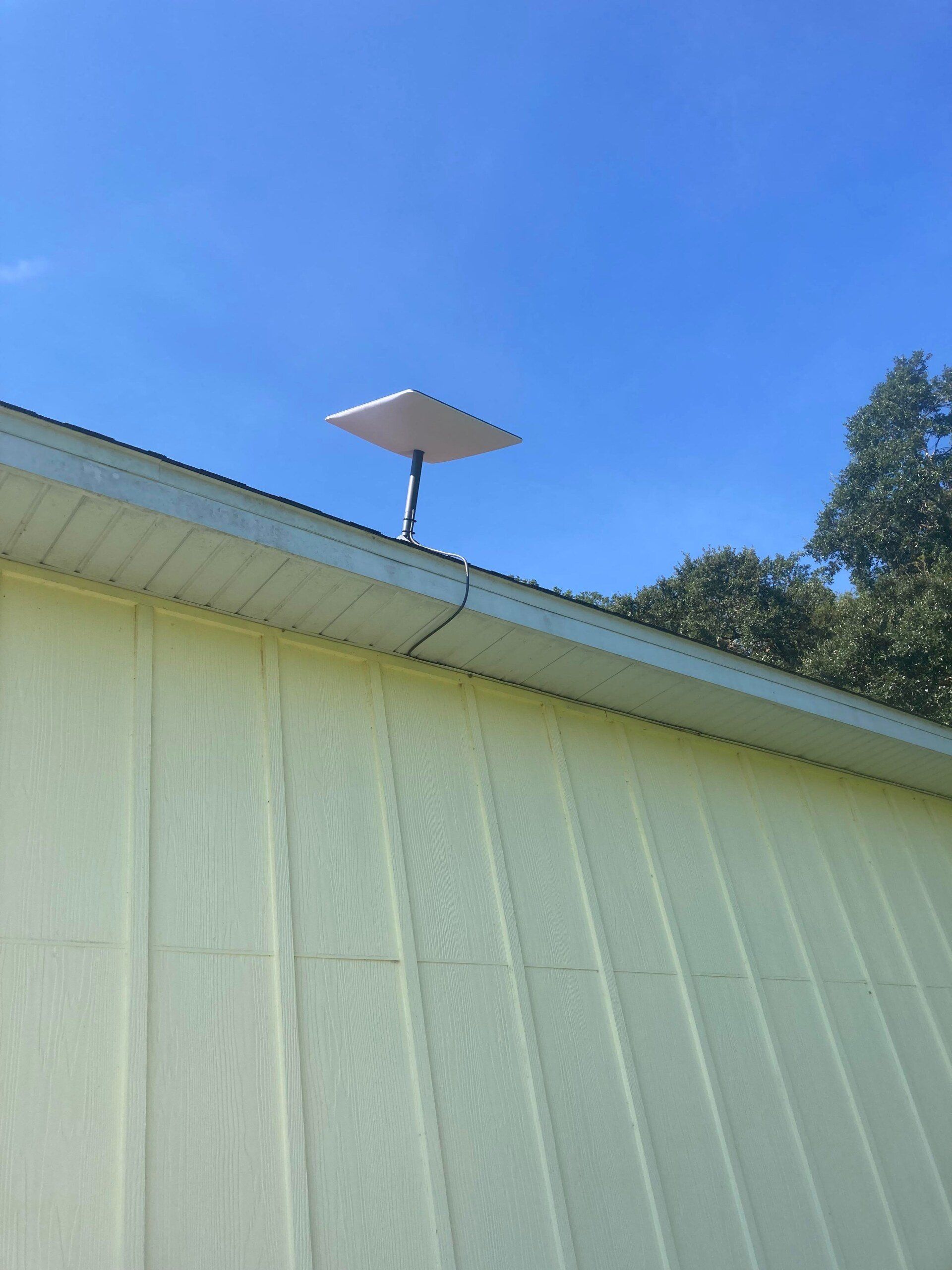 A square, white satellite dish mounted on a metal pole atop a yellow-sided building against a blue sky.