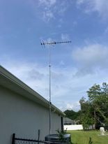 A tall TV antenna mounted on a pole beside a house, against a partly cloudy blue sky.