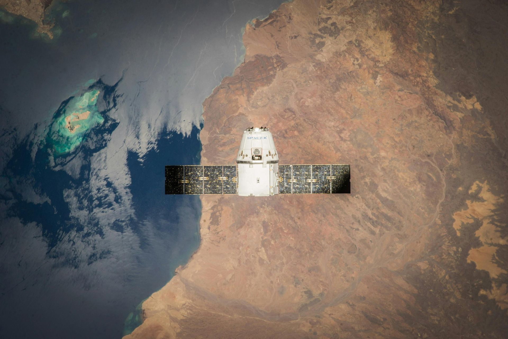 SpaceX Dragon spacecraft in orbit over Earth, solar panels extended. Dark blue ocean, brown landmass.