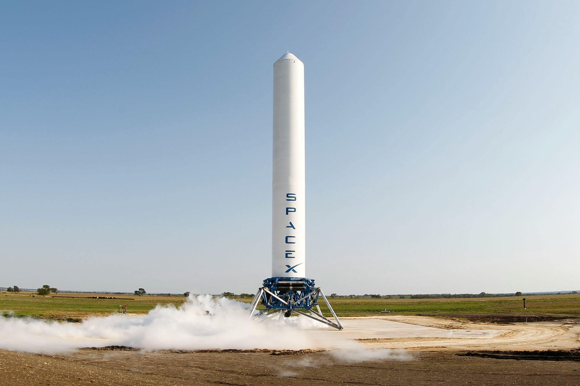 White SpaceX rocket on a launch pad, smoke rising, blue sky, and brown field.