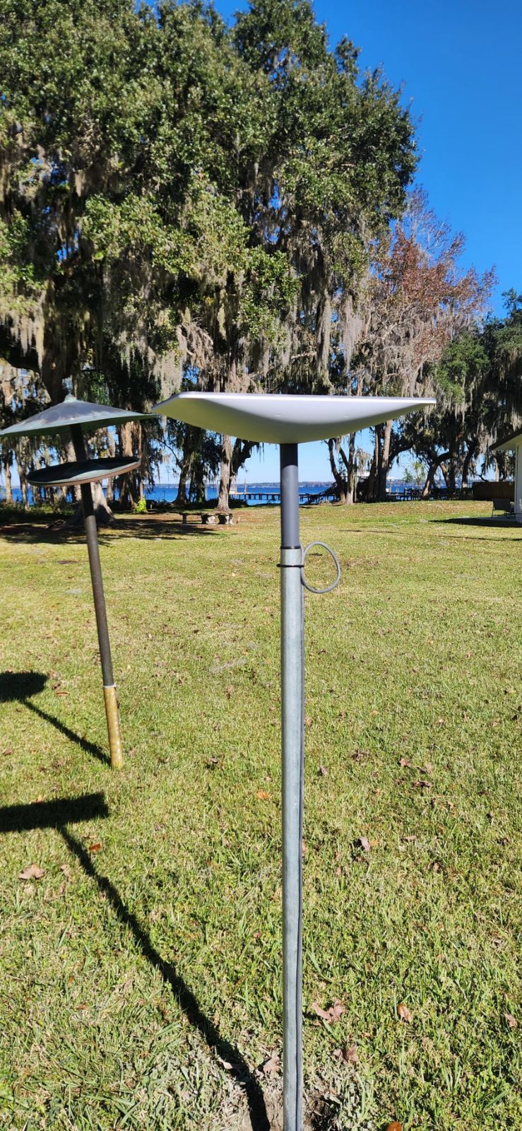 A metal birdbath on a pole in a grassy park, trees and water in the background, under a blue sky.