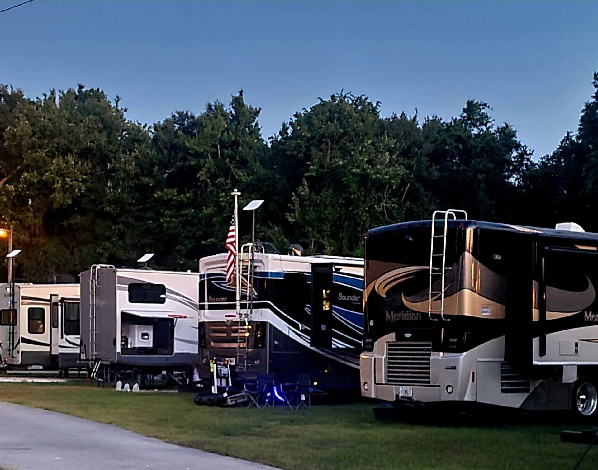 RVs parked at a campground, some with awnings and an American flag, at dusk. Trees in background.