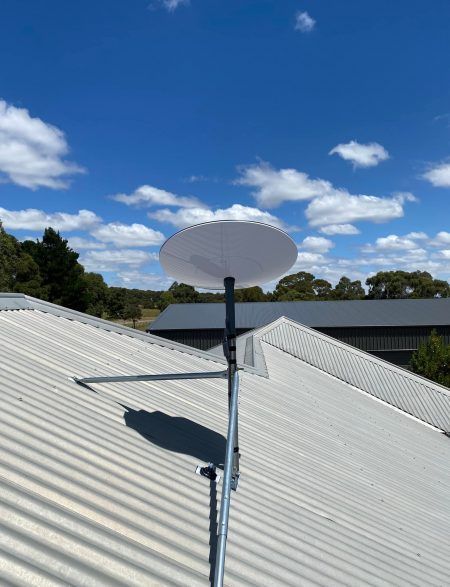 White satellite dish on a metal pole mounted on a corrugated metal roof against a blue sky with clouds.