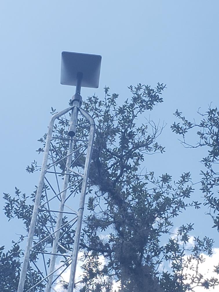 Metal tower with square top against a partly cloudy sky, behind a tree with green leaves.