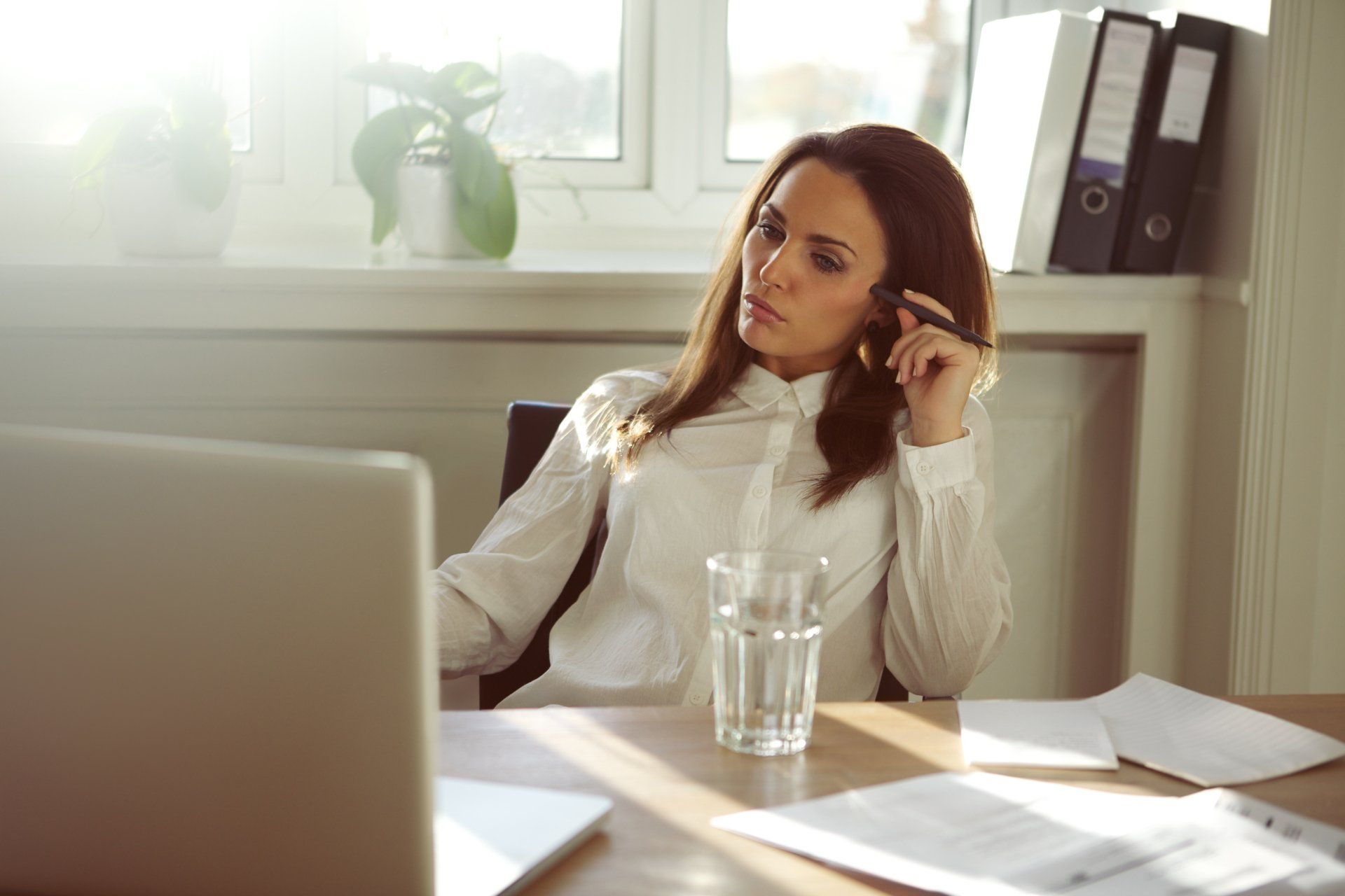 Woman in white shirt, seated at desk with laptop, pondering, holding pen near face, near window and papers.