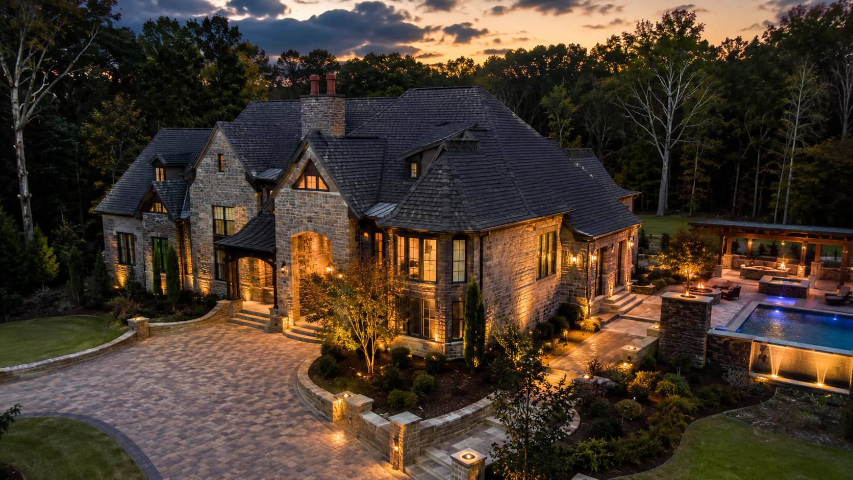 Luxury stone house at dusk with warm lights, curved driveway, and a lit backyard pool surrounded by trees