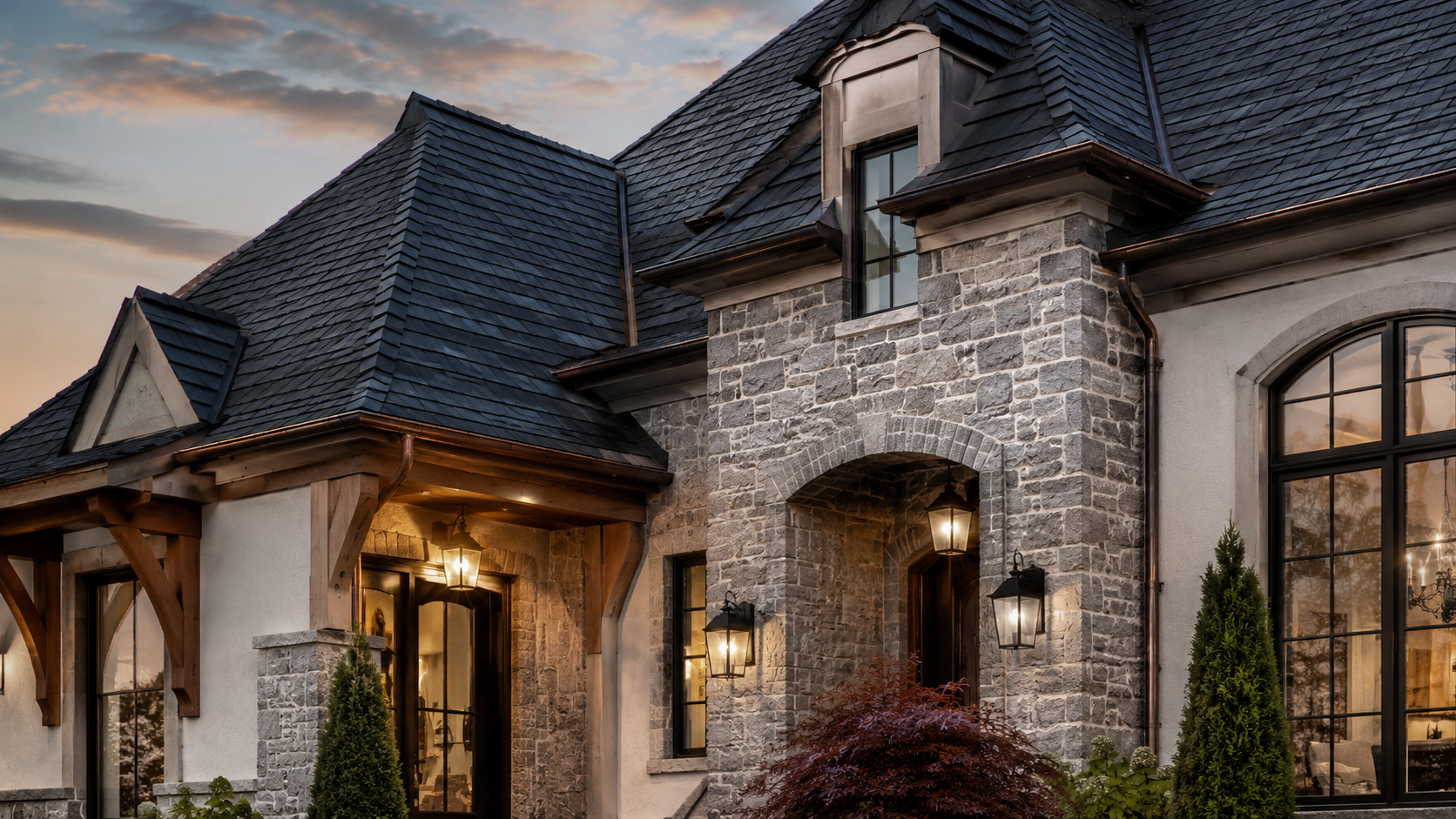 Large stone-and-wood house exterior at dusk with lit porch, dark roof, and arched windows