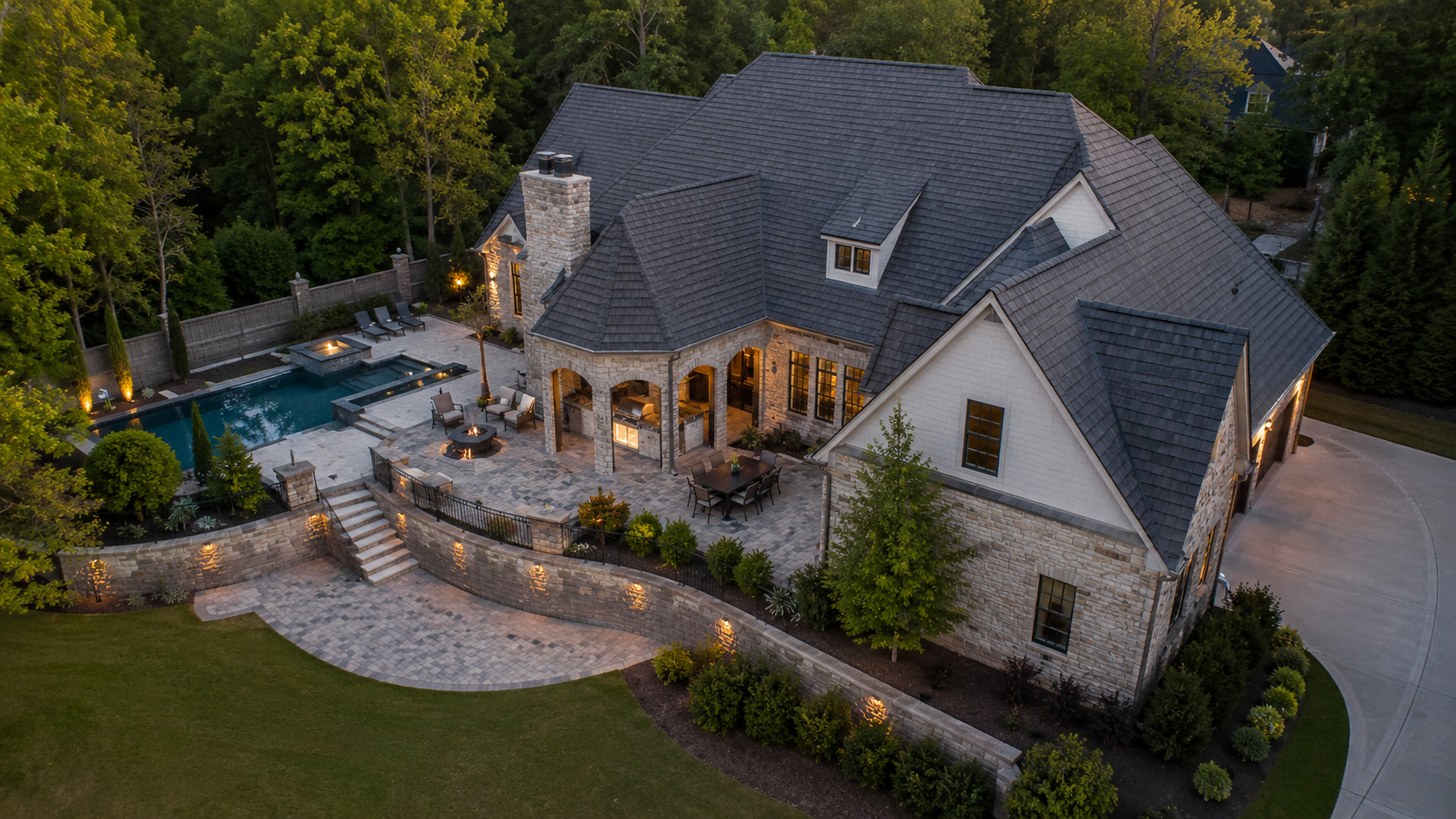 Aerial view of a large stone house with lit patio, pool, and landscaped garden at dusk