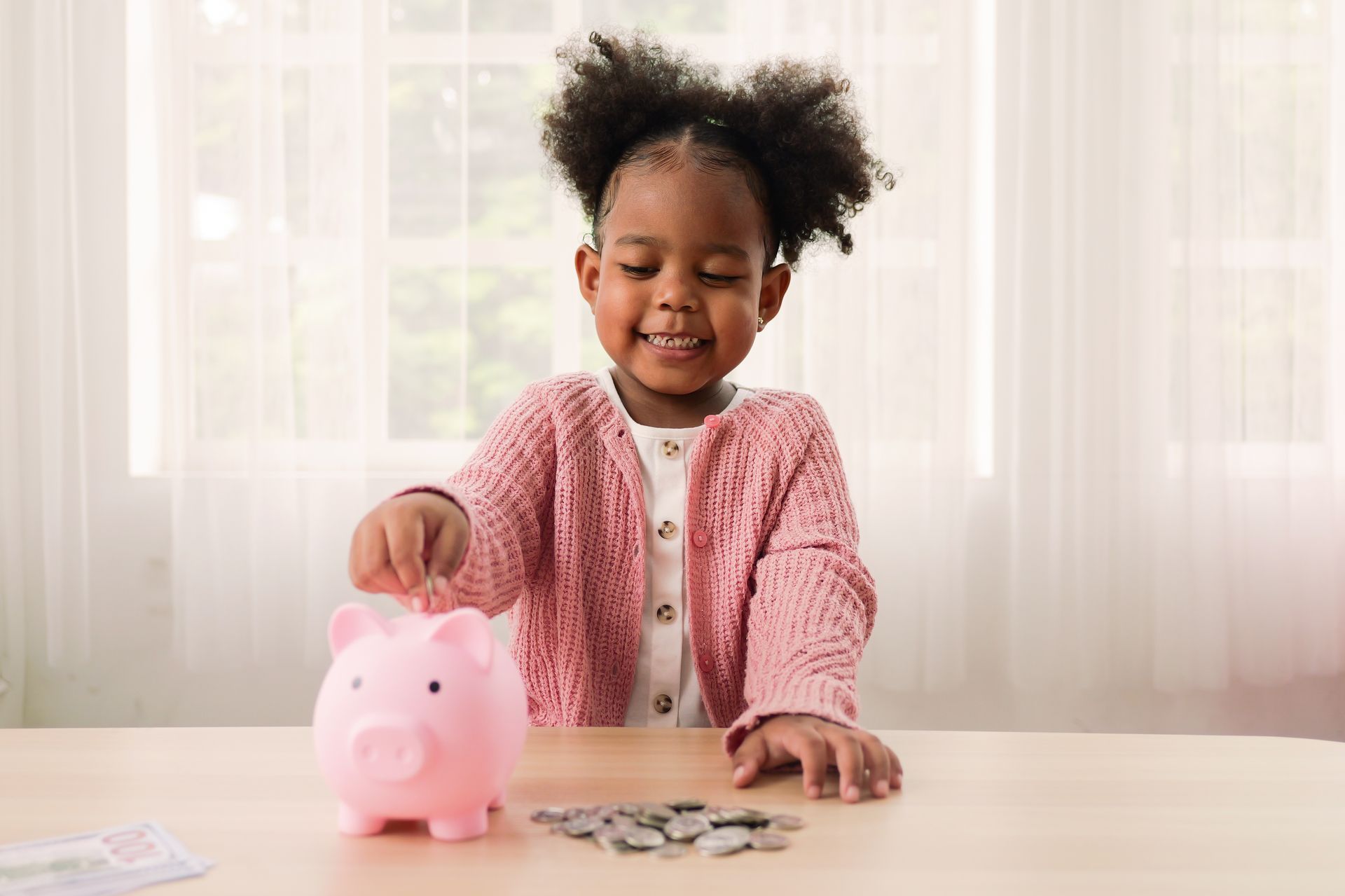 Child smiling while placing coins into a pink piggy bank at a table