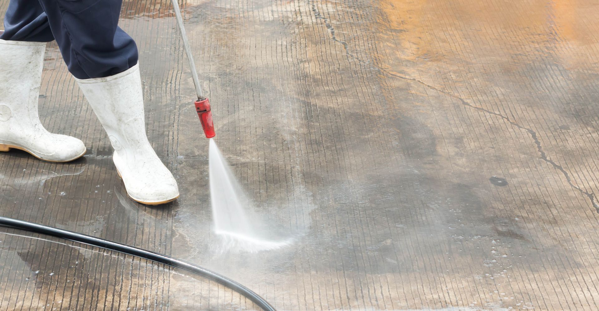 Person pressure-washing a concrete surface with a red spray wand and white boots.
