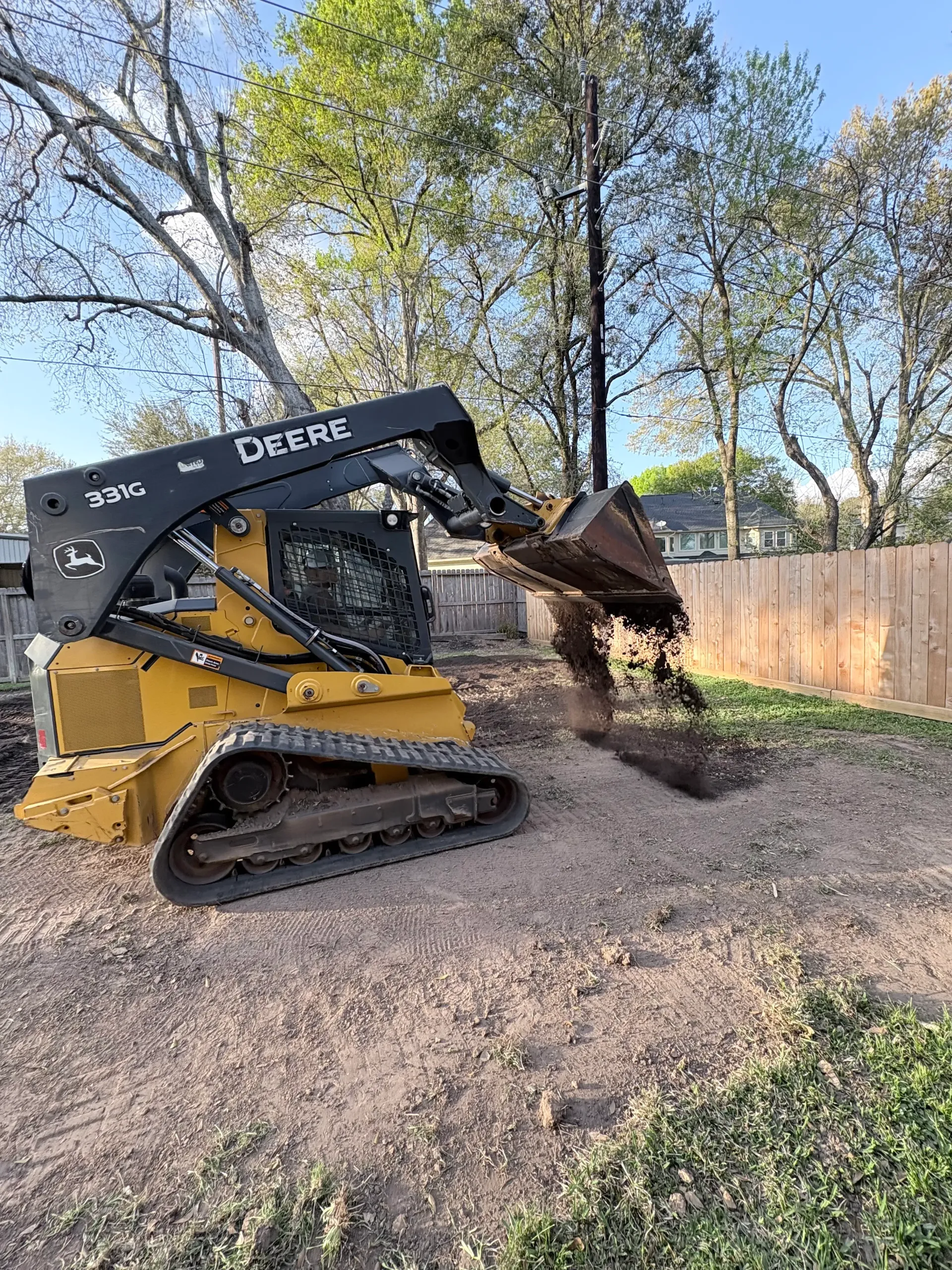 A yellow John Deere compact track loader scoops dirt in a residential yard near a wooden fence and trees.