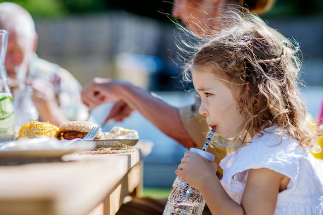 Girl drinks from a bottle with a straw at an outdoor table; other people and food are visible.