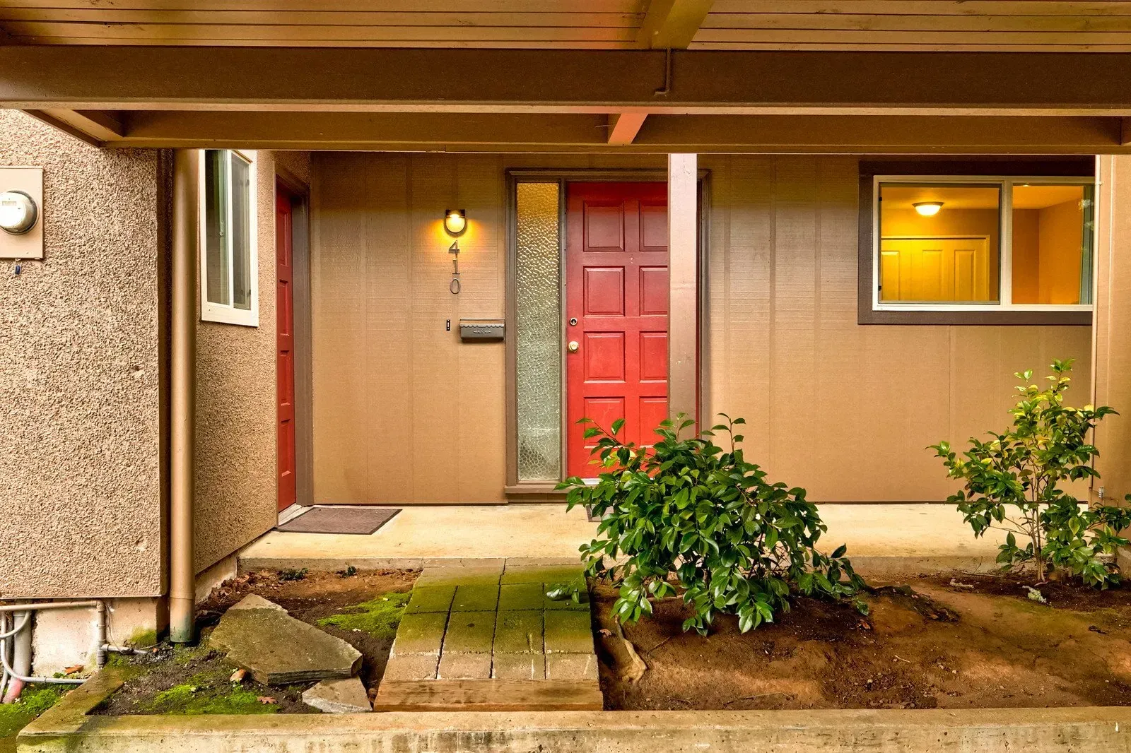 Red door of a home under a brown overhang, with a small garden in front.