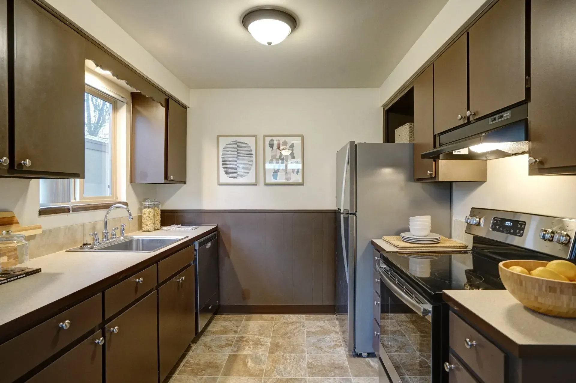 Dark brown kitchen cabinets and appliances in a narrow kitchen with light-colored countertops.