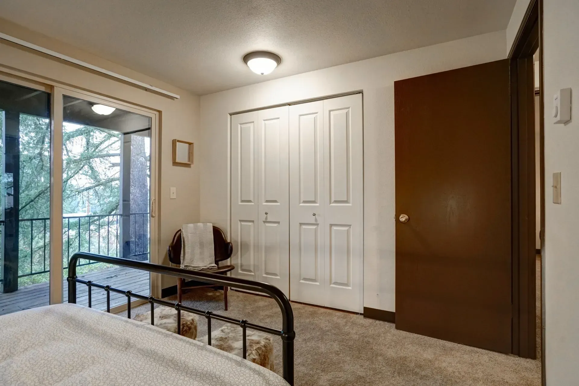 Bedroom with sliding glass door, white closet doors, dark brown door, chair with blanket, and black metal bed frame.