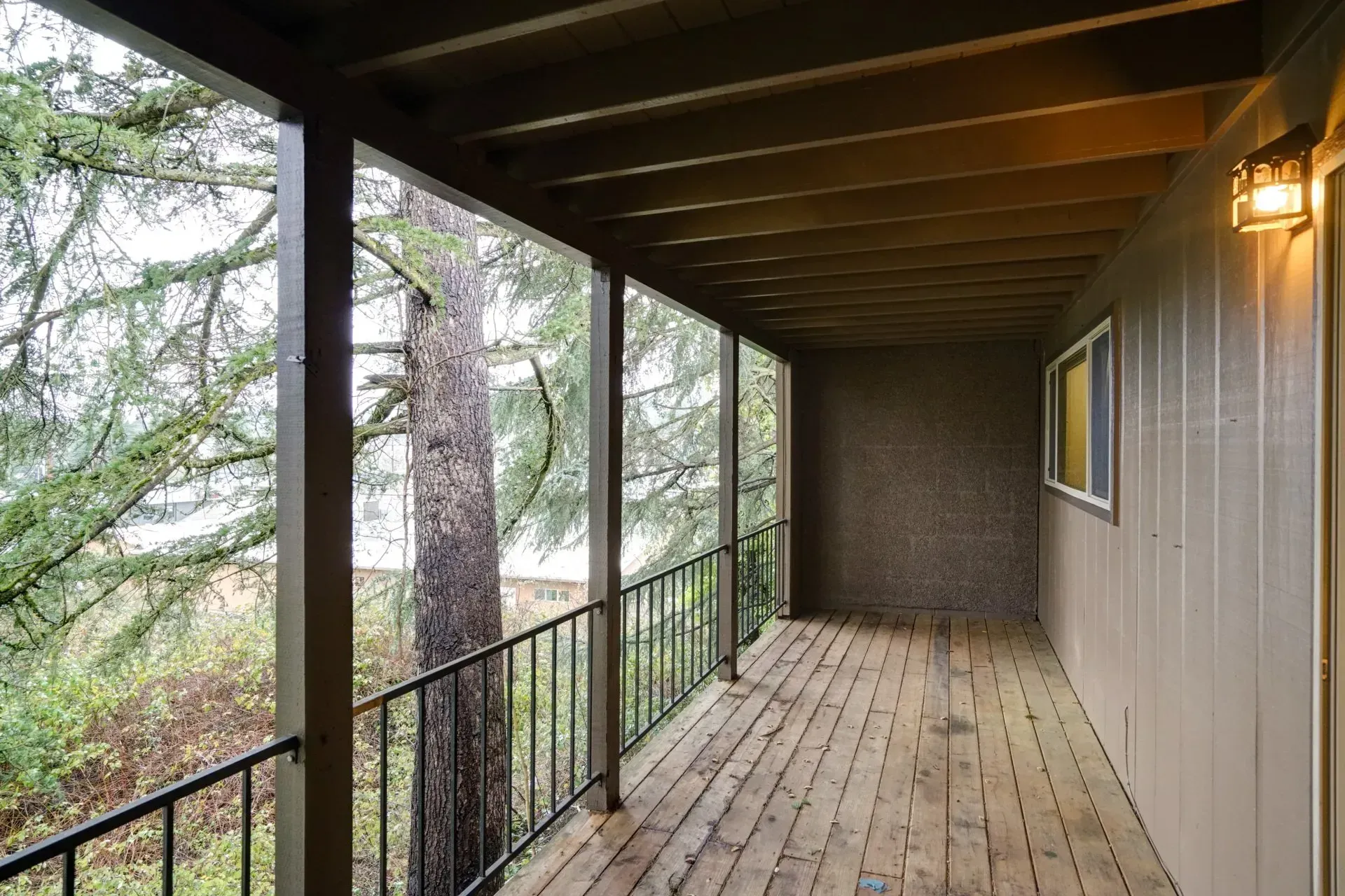 Covered wooden deck with railing, tree view. Brown wooden planks, beige siding, warm light, overcast day.
