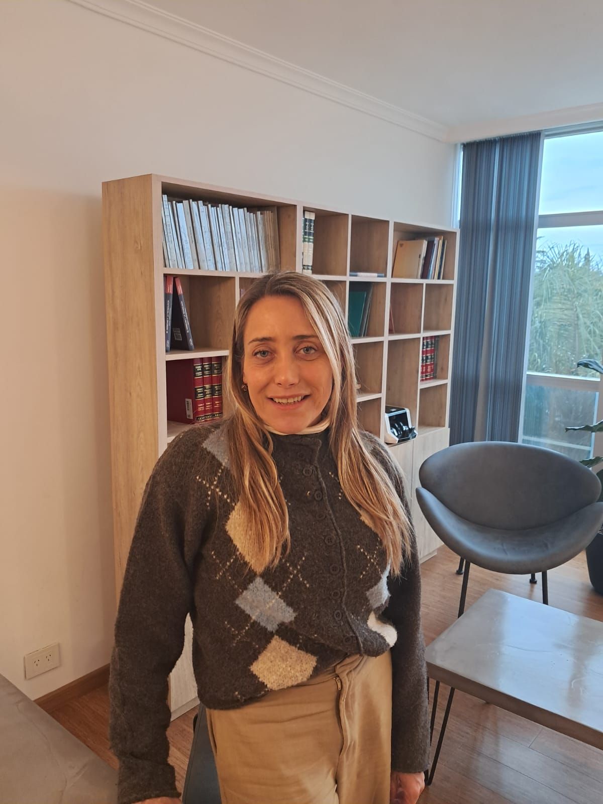 Persona de pie en una luminosa sala de estar con una estantería, una silla y vistas a la ventana.