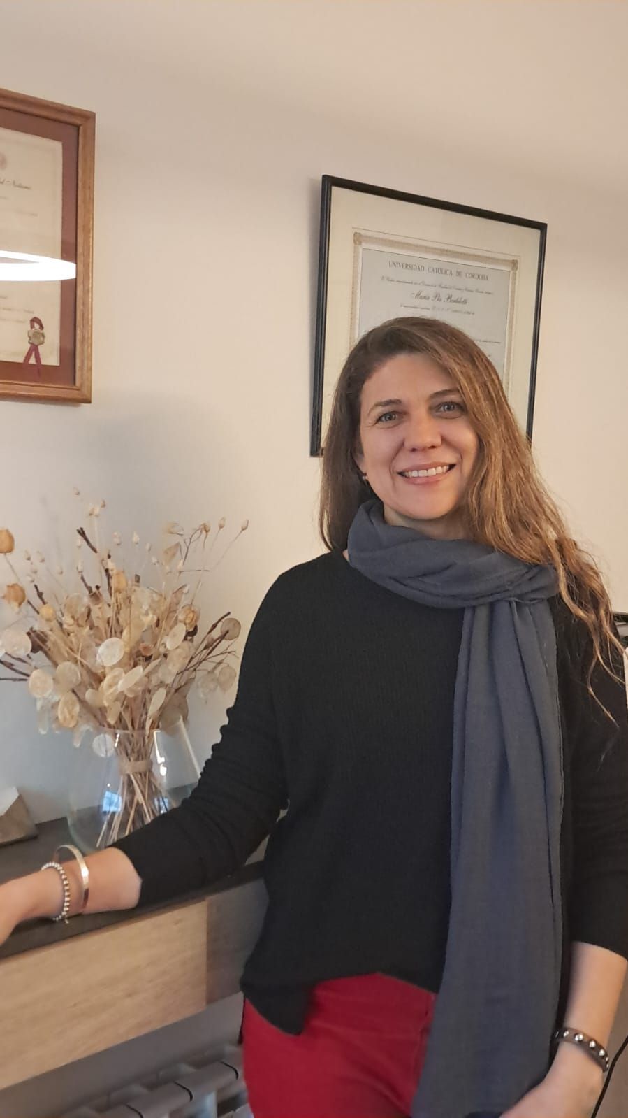 Mujer de pie en el interior de una casa, junto a una estantería, con una blusa negra y una bufanda gris, sonriendo a la cámara.