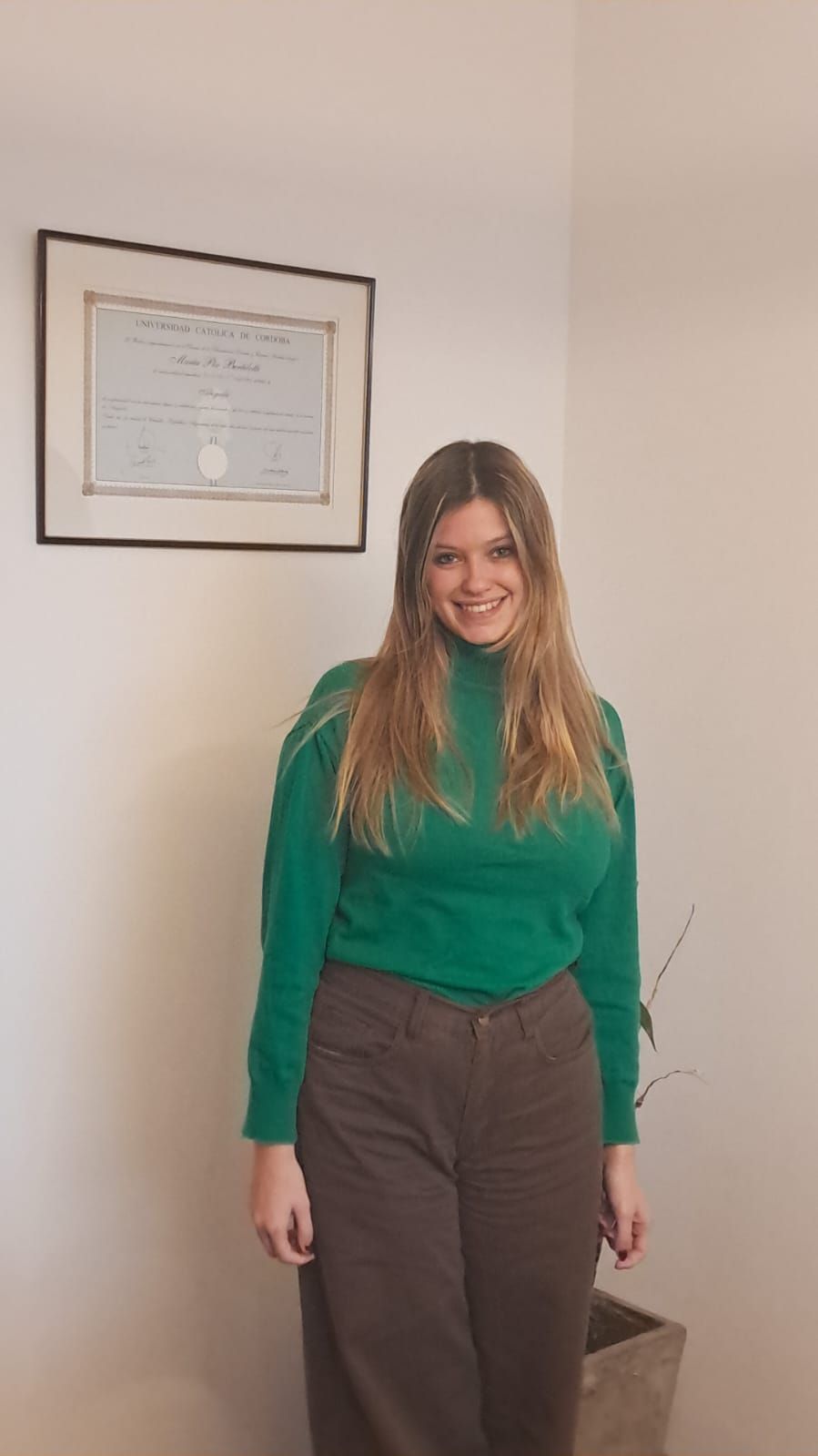 Mujer con blusa verde y pantalones oscuros, de pie en el interior de un edificio, sonriendo junto a un certificado enmarcado.