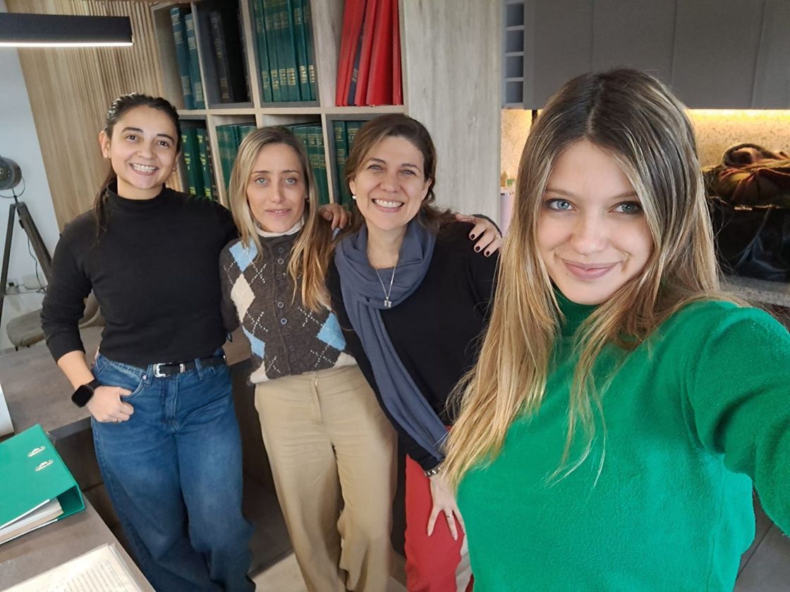 Cuatro personas de pie en el interior, sonriendo y posando juntas cerca de una ventana y una mesa.